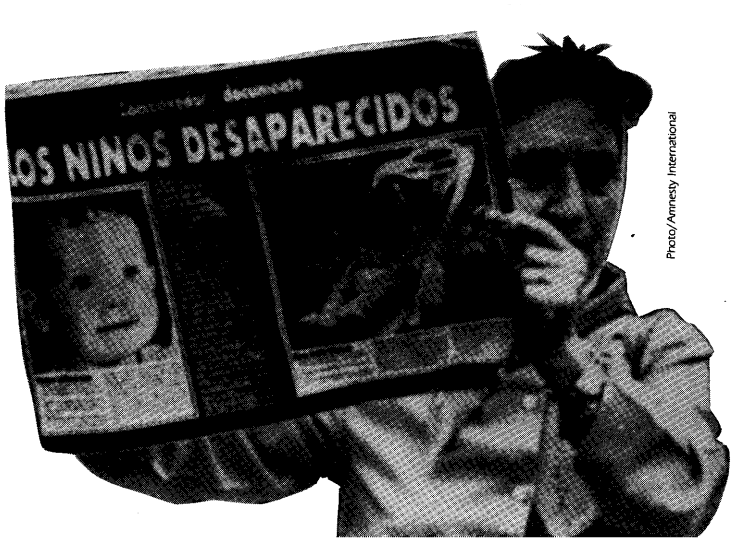 A grandmother in a coat holds up a sign that says "Los Ninos Desaparecidos"